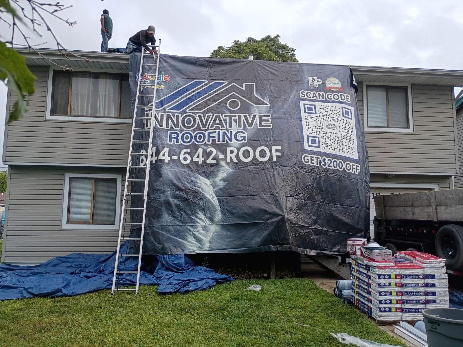 Gray two-story home mid replacement with Innovative Roofing banner, crew on the roof, and Timberline HD shingles staged