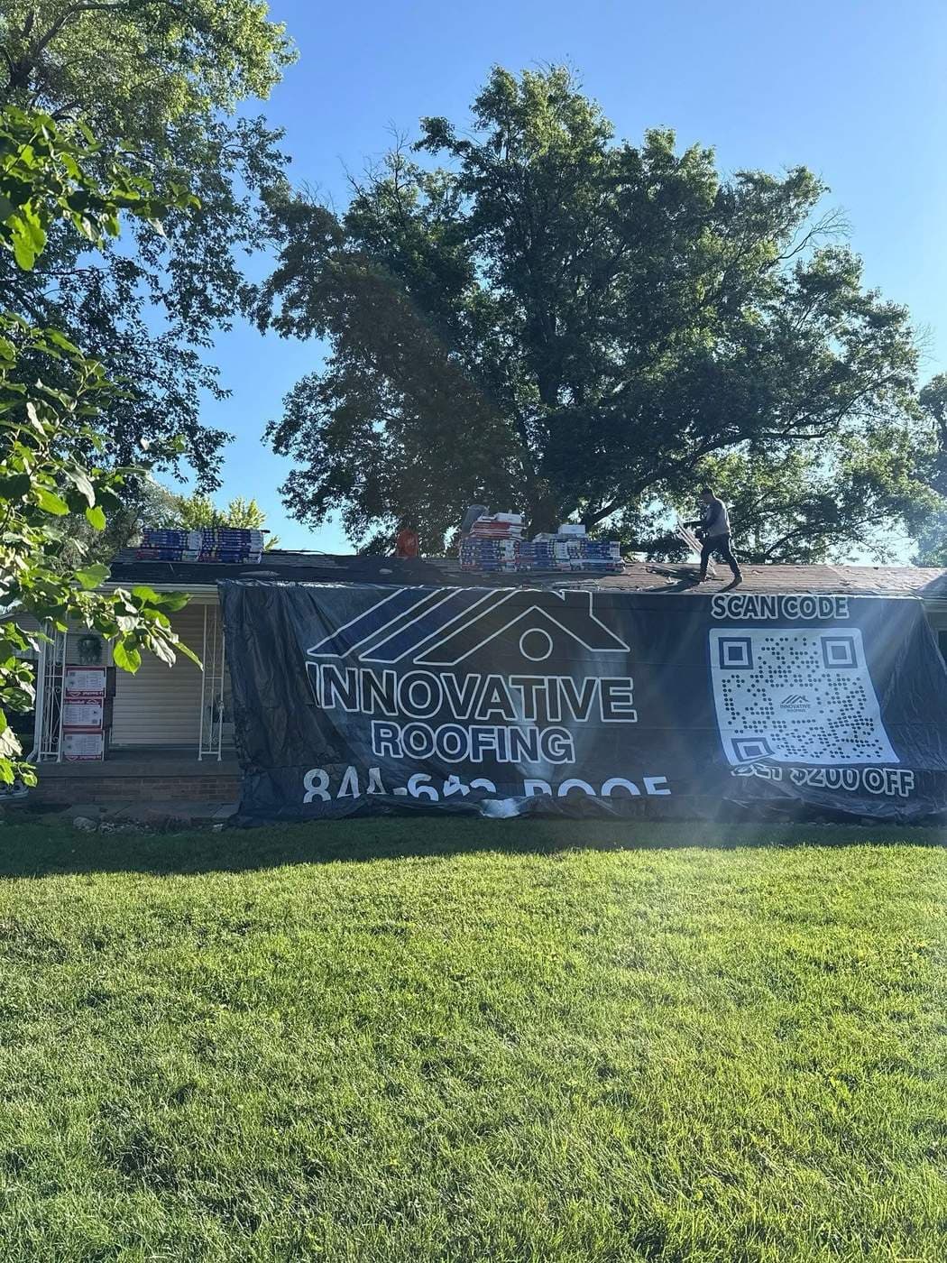 Single-story home with Innovative Roofing promotional banner and shingle bundles staged along the ridge