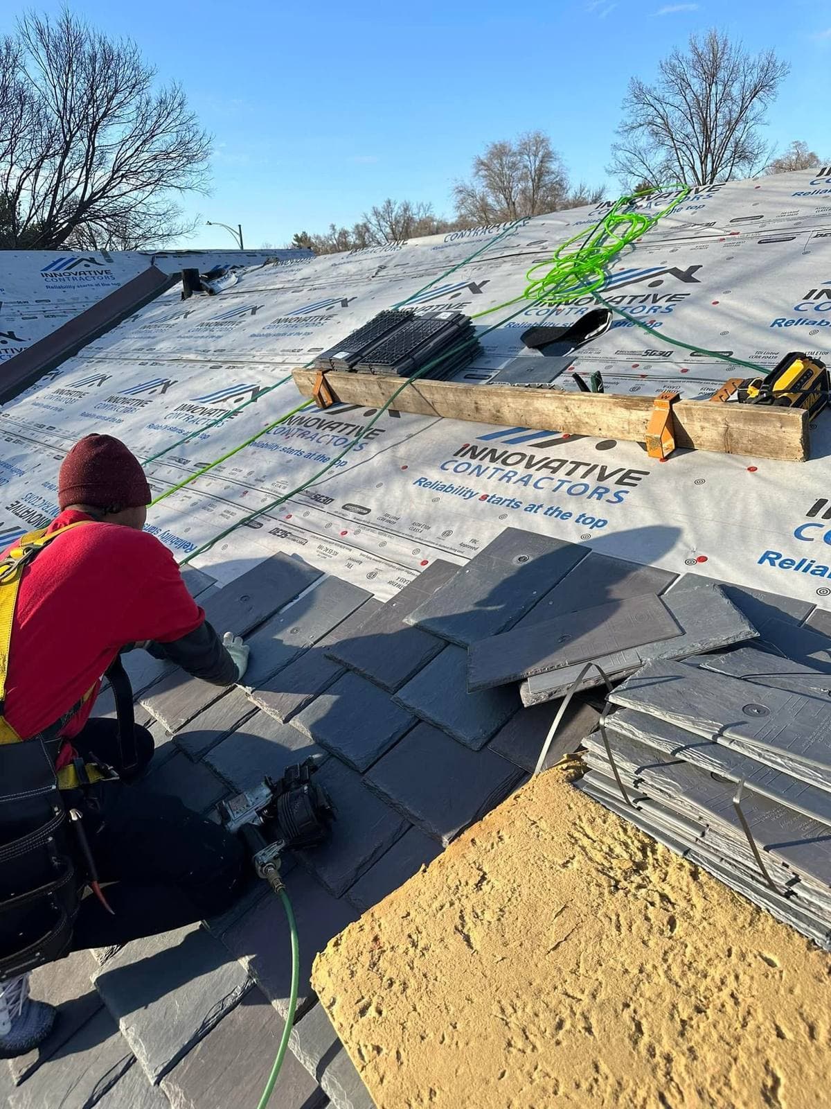 Roofer in a safety harness nailing dark slate-style shingles over Innovative Contractors branded underlayment