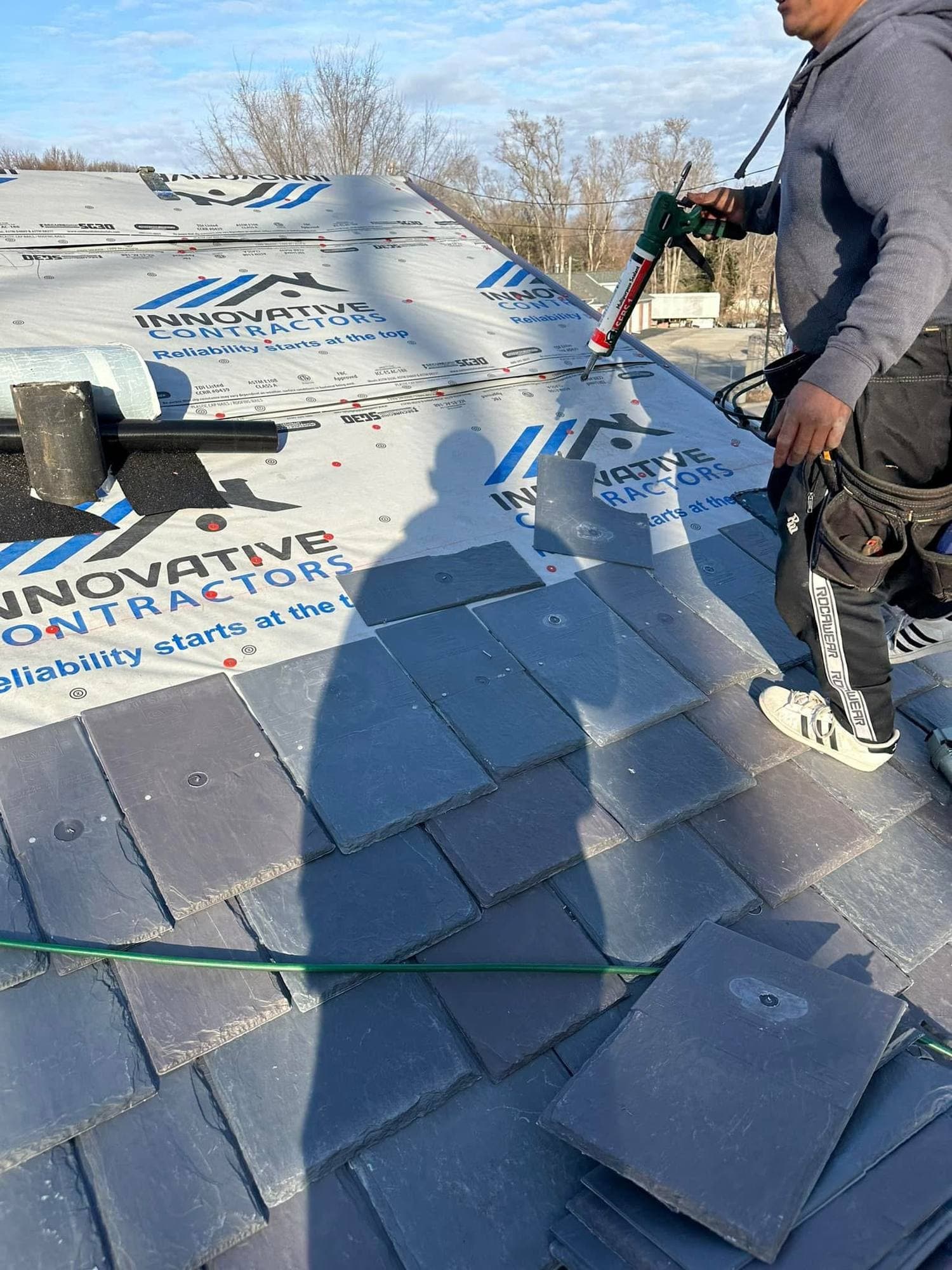 Roofer applying sealant to a dark slate-style shingle beside Innovative Contractors branded underlayment