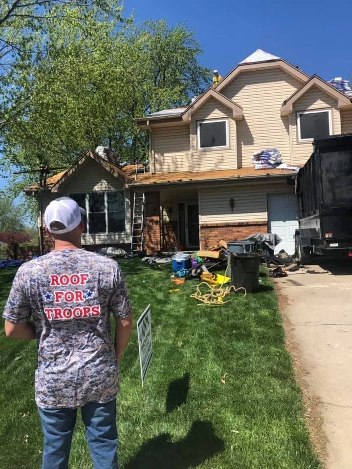 Team member in a Roof for Troops shirt watching a two-story home mid roof replacement with materials staged on the deck