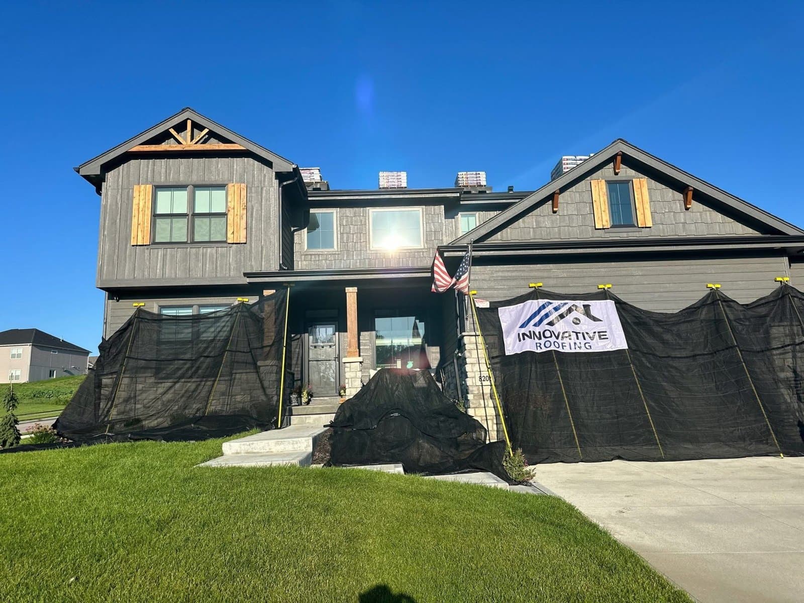 Modern gray two-story home with Innovative Roofing banner on debris netting and new shingle bundles staged on the roof ridge
