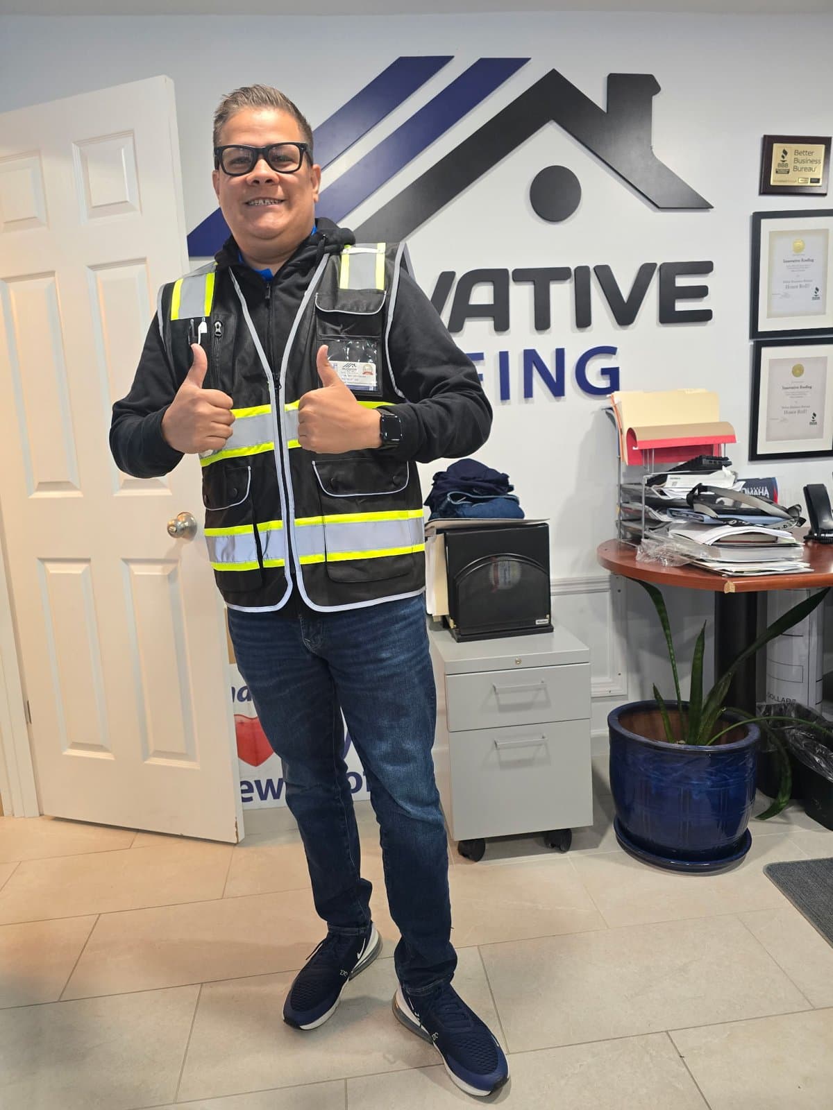 Innovative Roofing team member in a safety vest giving two thumbs up in the office in front of the company logo wall
