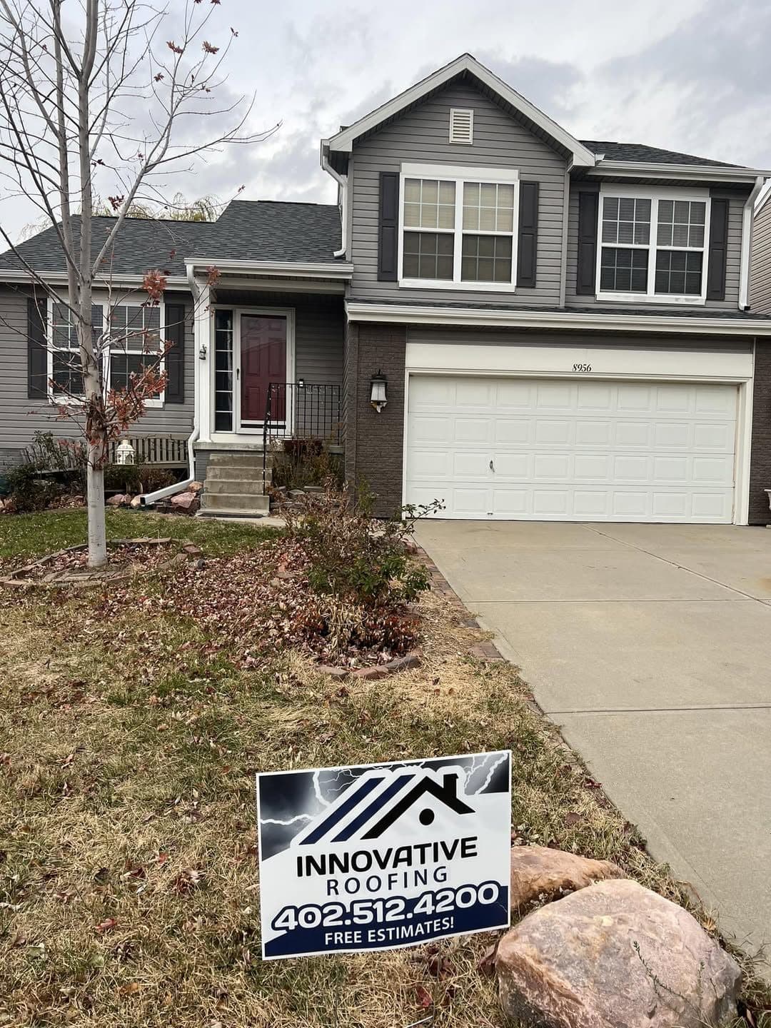 Two-story home with gray siding, brick base, and Innovative Roofing yard sign on the front lawn