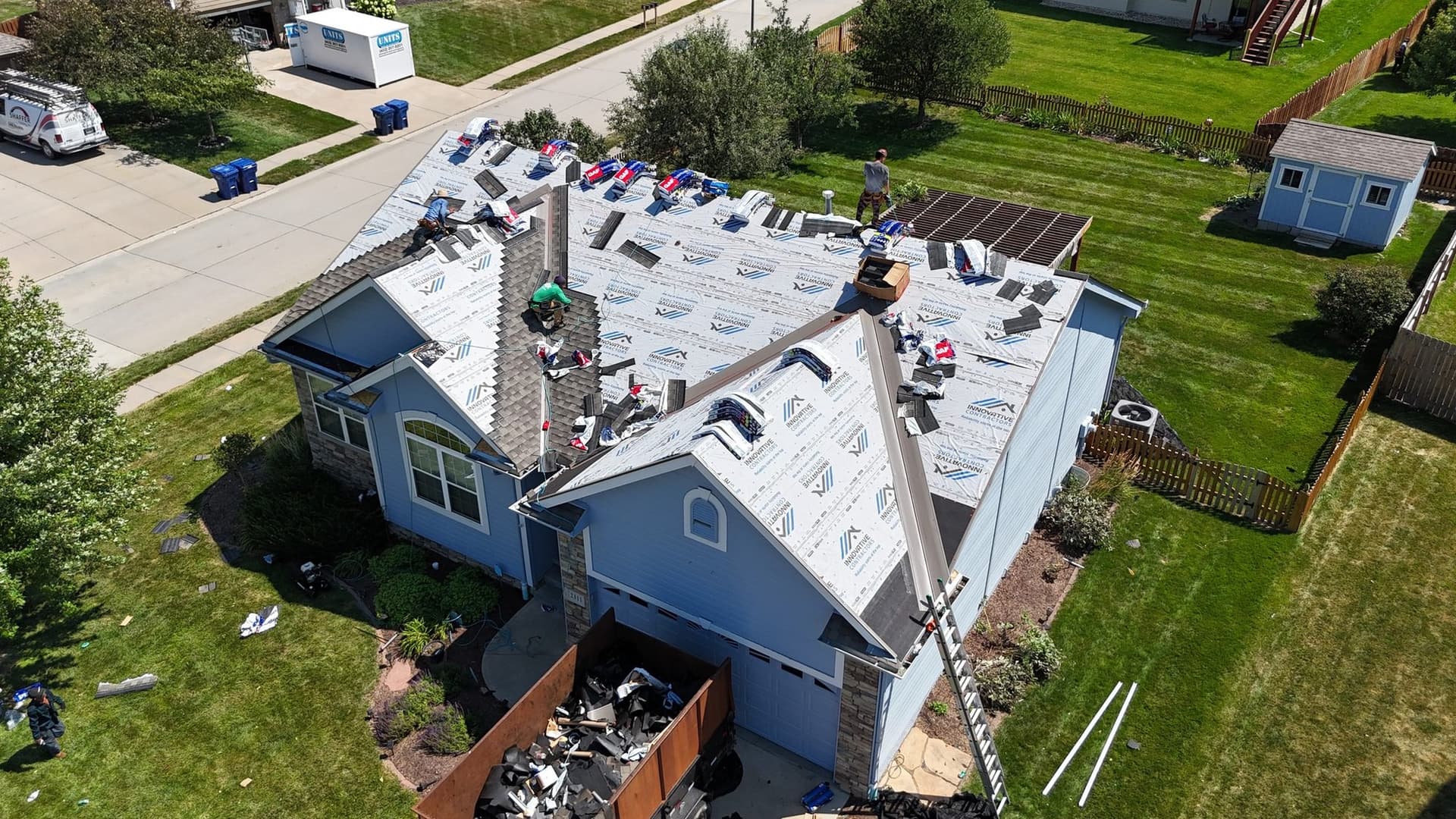 Aerial drone view of a blue two-story home during an Innovative Roofing replacement with branded underlayment and a debris dumpster