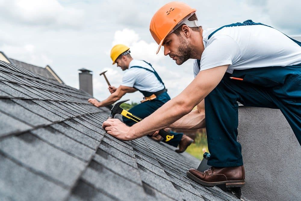 Two harnessed roofers in hard hats and safety vests working a steep shingle roof